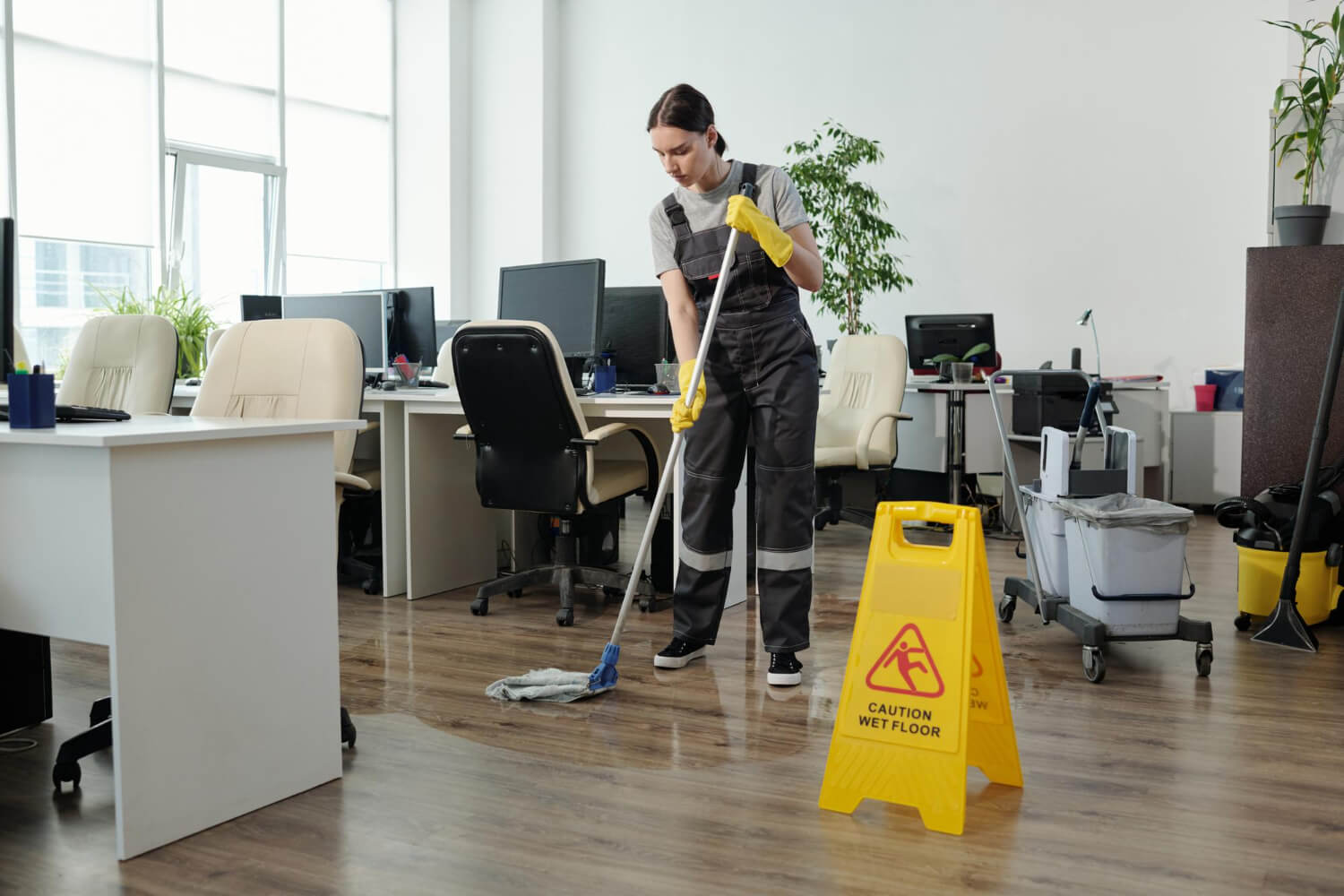young female cleaner in yellow protective gloves and grey uniform mopping office floor