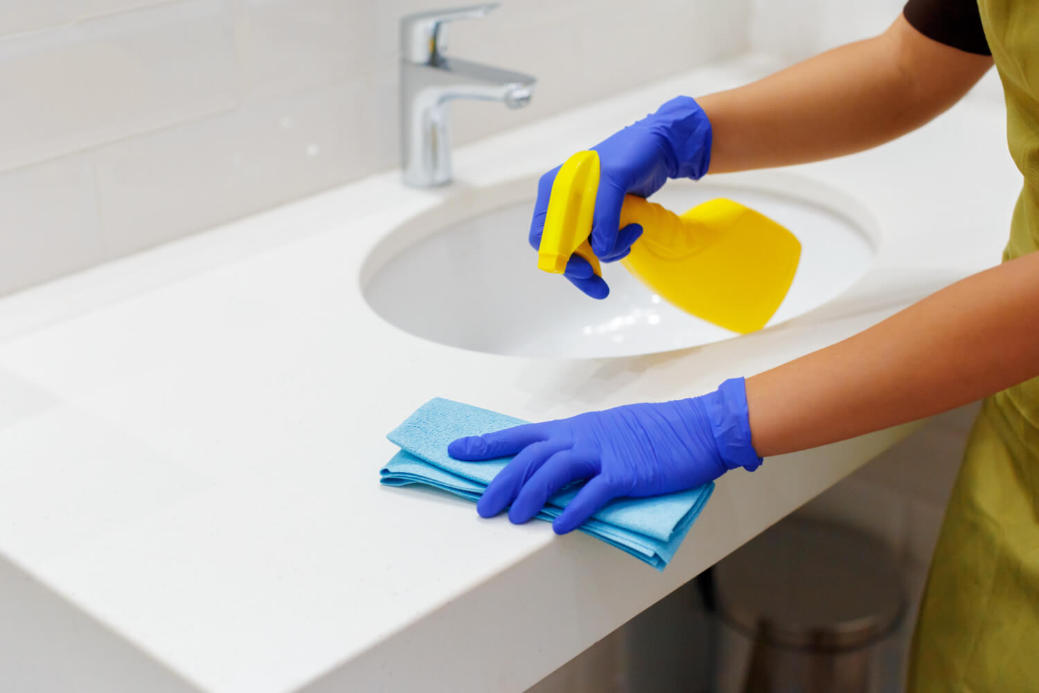 close up cleaners hands in blue gloves cleaning white bathroom sik with blue cloth and yellow spray