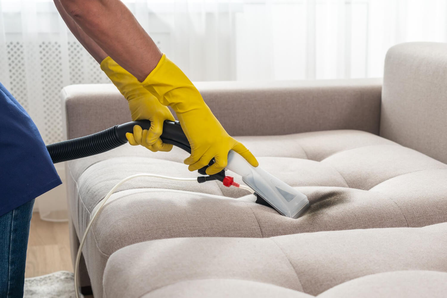 close up cleaners hands in yellow protective gloves holding professional vacuum cleaner while cleaning grey sofa