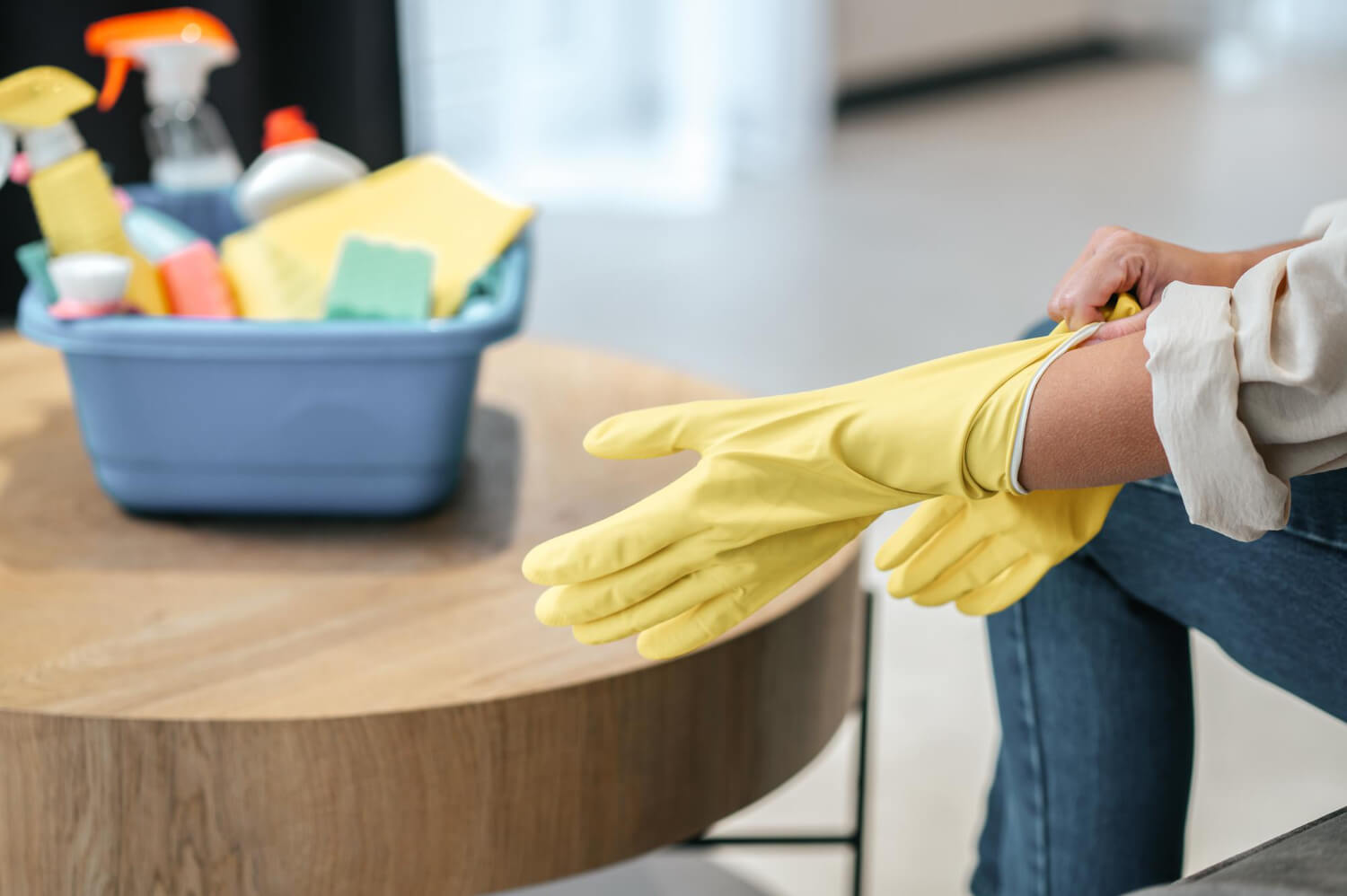 close up females hands wearing yellow cleaning gloves with bucket with cleaning equipment on table next to her