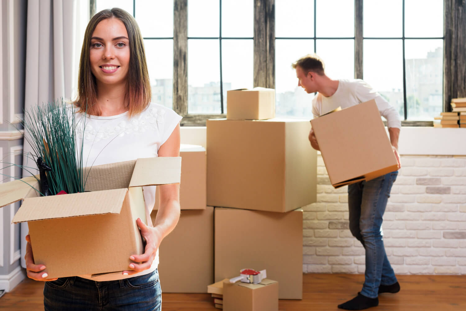 woman holding cardboard box with plants and man behind them holding closed moving boxe with staffed boxes around him