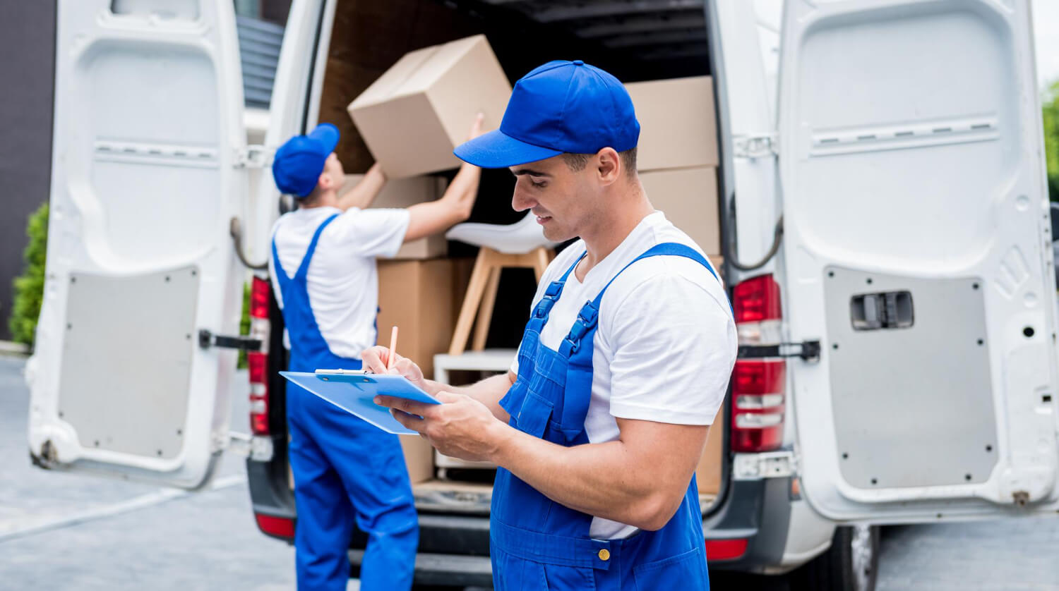 two removal company movers in the back of a white moving back one loading boxes and the other checking the list of things in notebook