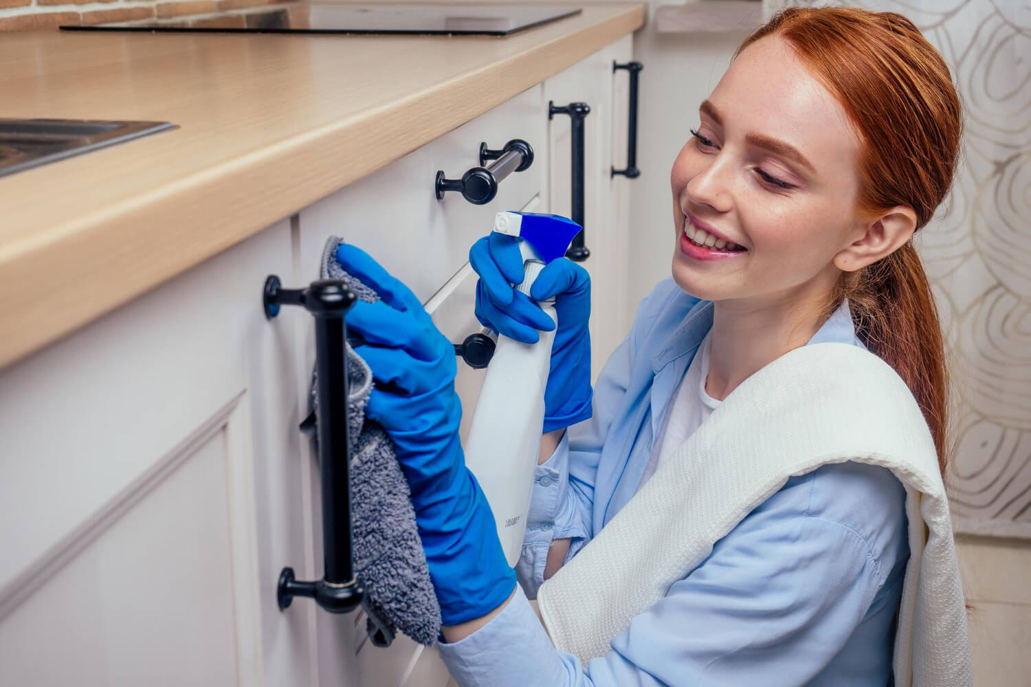 red haired woman close up smilling while cleaning kitchen with blue gloves and holding cleaning spray and grey cloth