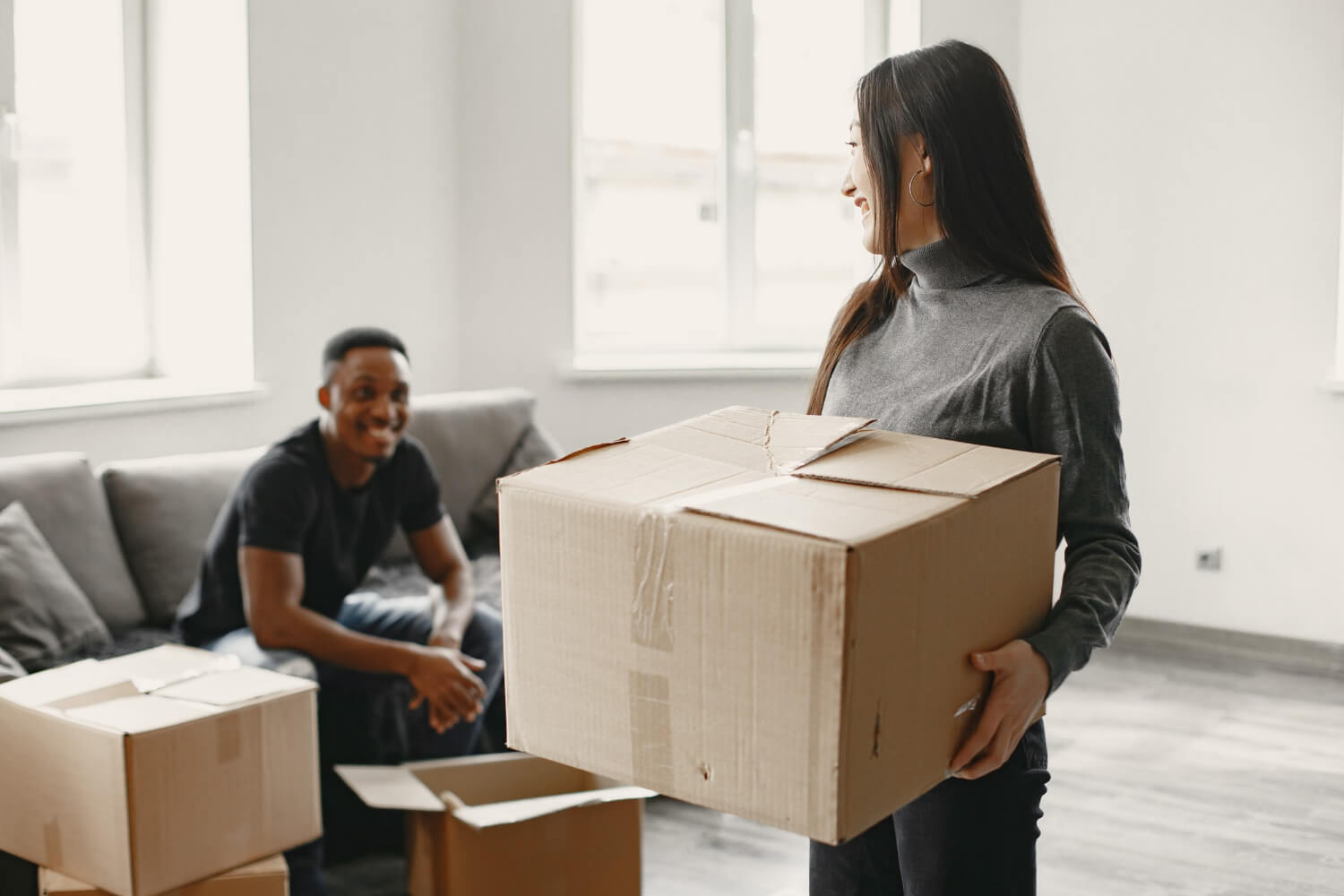 young man sitting on sofa with moving boxes around him smiling to woman who holds moving box for relocation