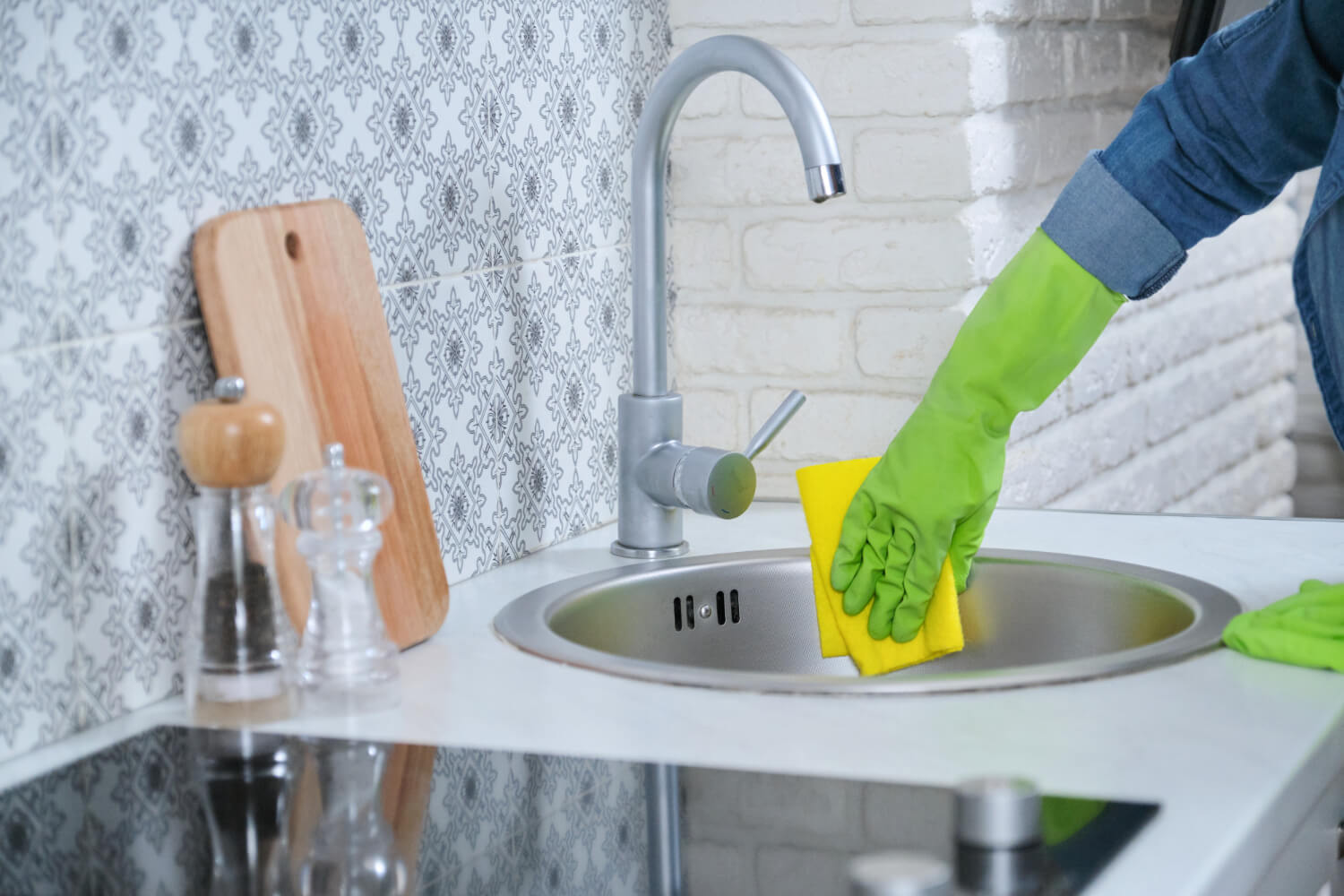close up cleaners hands in jean shirt and green protective gloves cleaning kitchen sink with yellow cloth