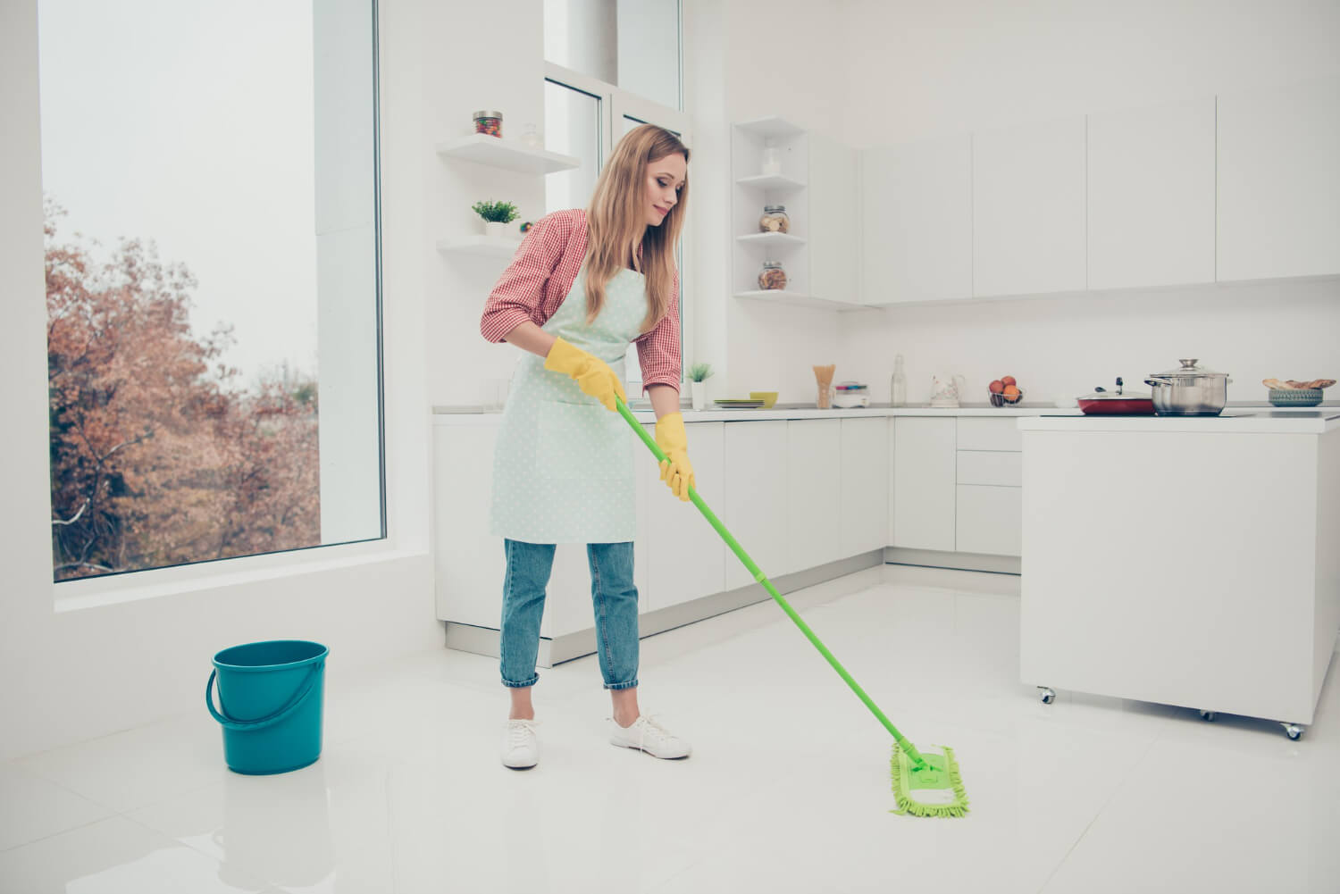 young blonde woman in jeans mopping white kitchen floor with green mop and blue bucket next to her