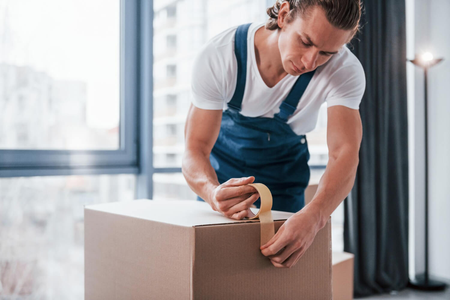 young male mover with blue uniform works indoors room and closing moving box with tape