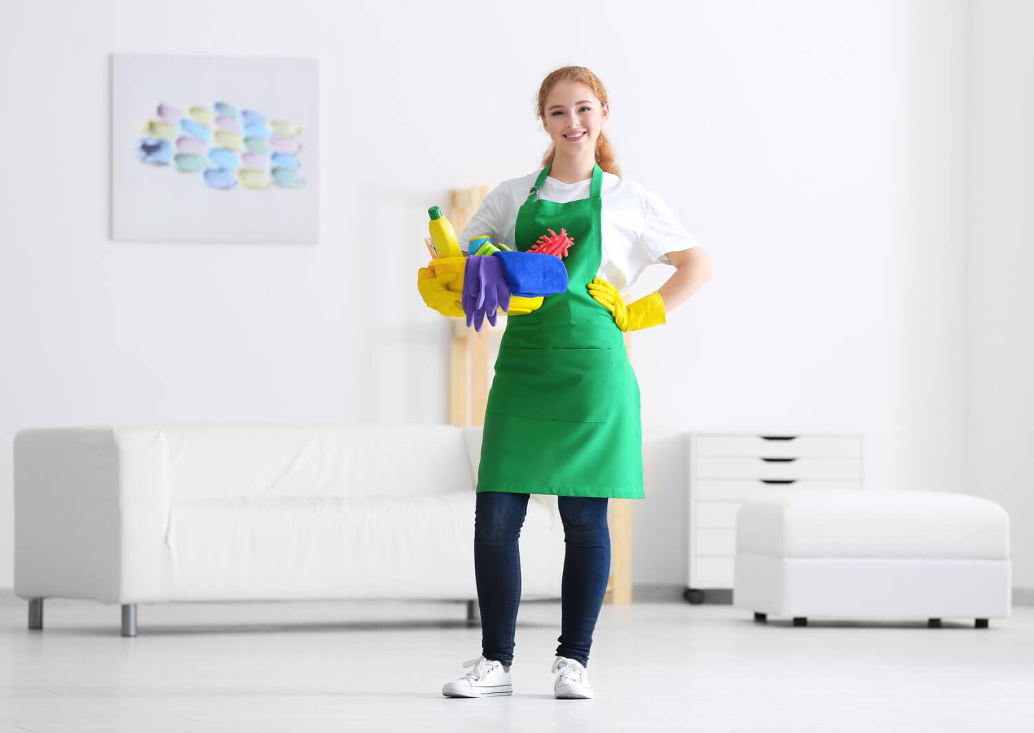 happy woman in green uniform and yellow gloves smiles at camera while holding yellow bucket with cleaning supplies