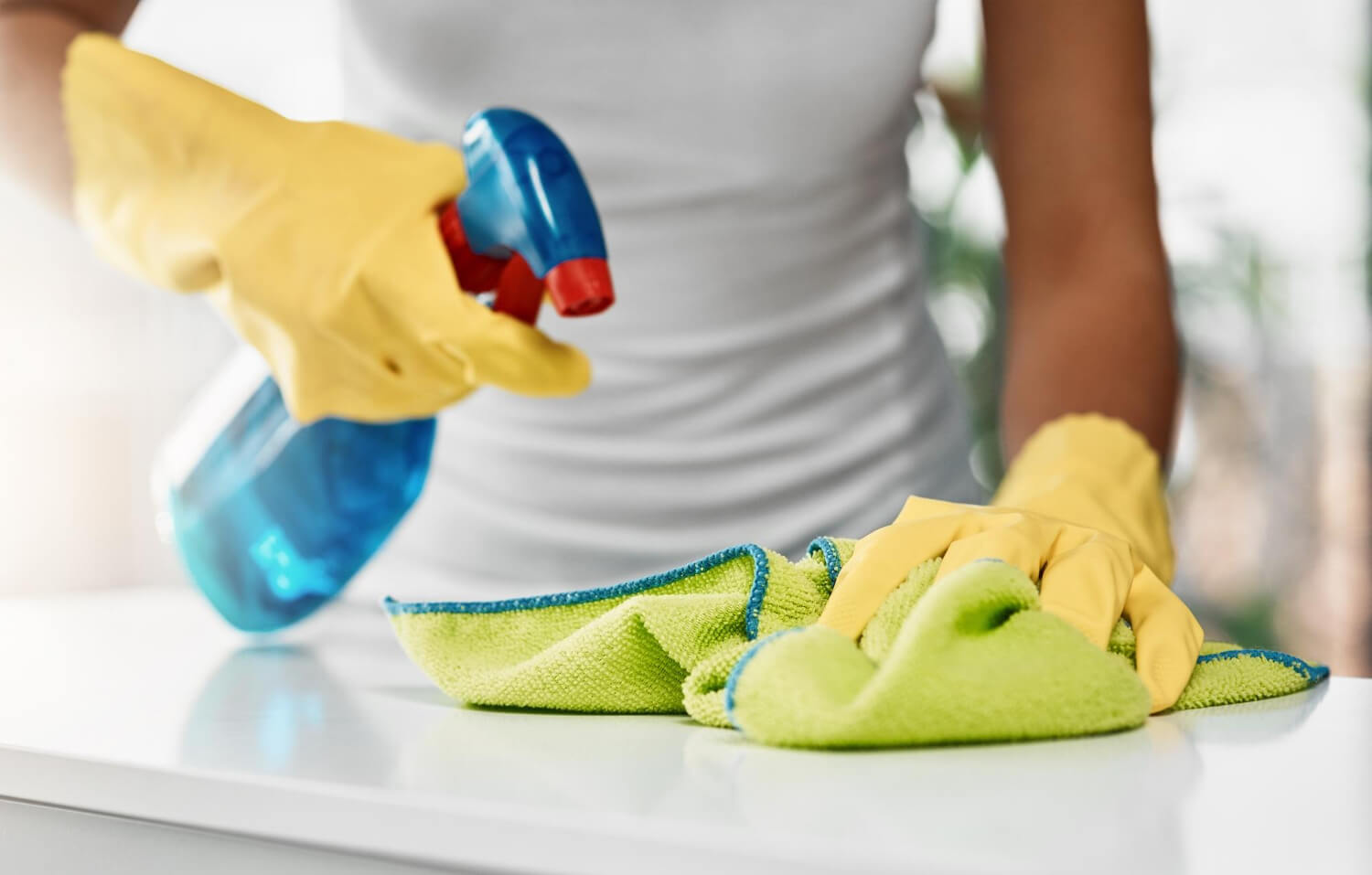 close up cropped female cleaners hands in yellow gloves cleaning white surface with blue spray and green cloth