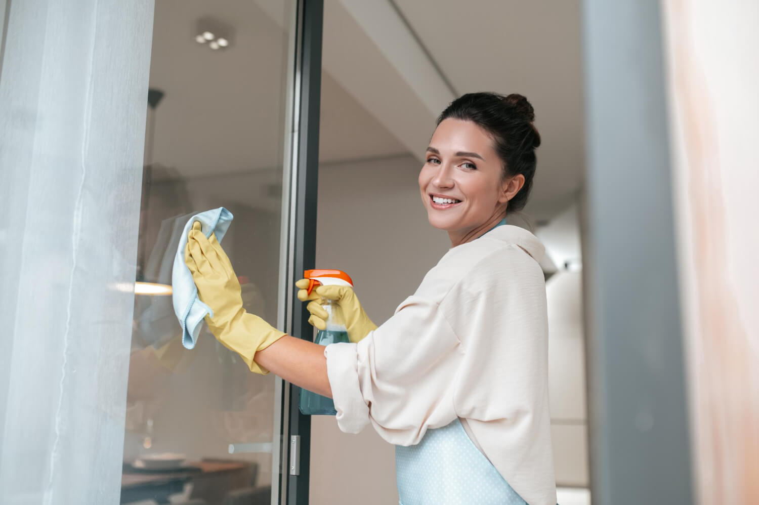 happy young woman in yellow gloves cleaning window with blue cloth and spray while smiling at camera