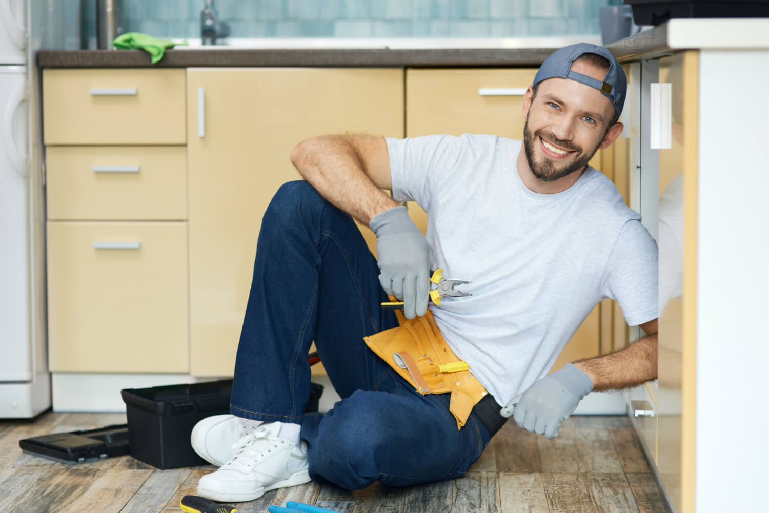 handyman sitting on kitchen floor wearing tool belt smiling on camera with open kitchen cabinet