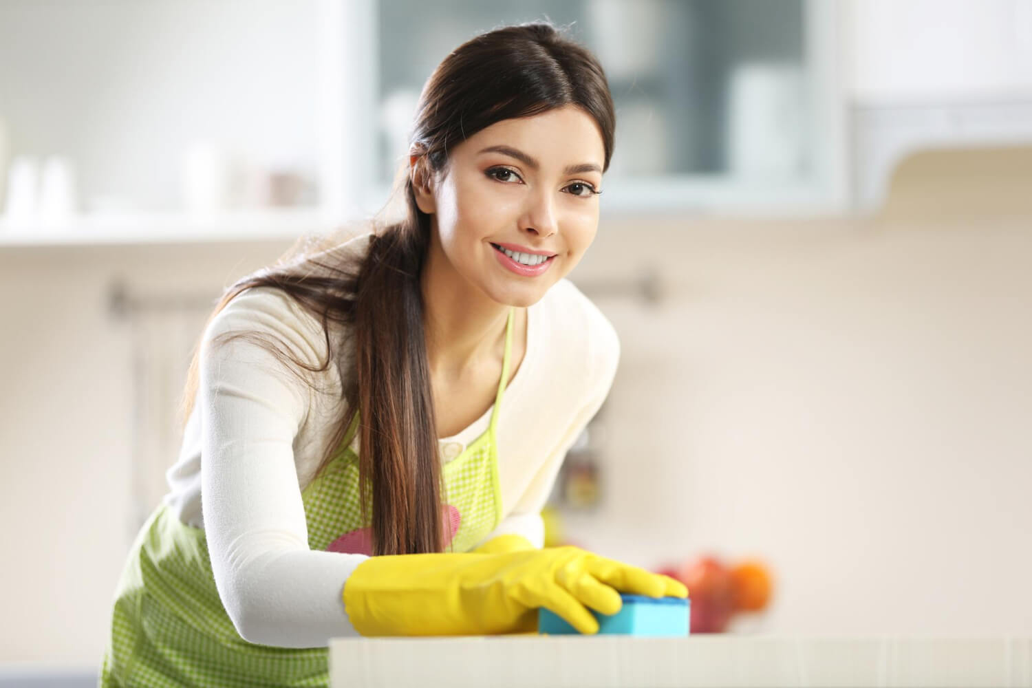 close up young woman in yellow protective gloves cleaning appliance with blue sponge while smilling at camera