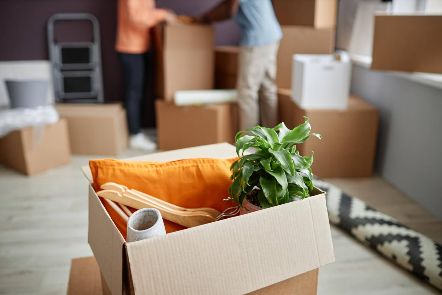 top view of open moving box filled with home belongings and blurred homeowners in the back packing things