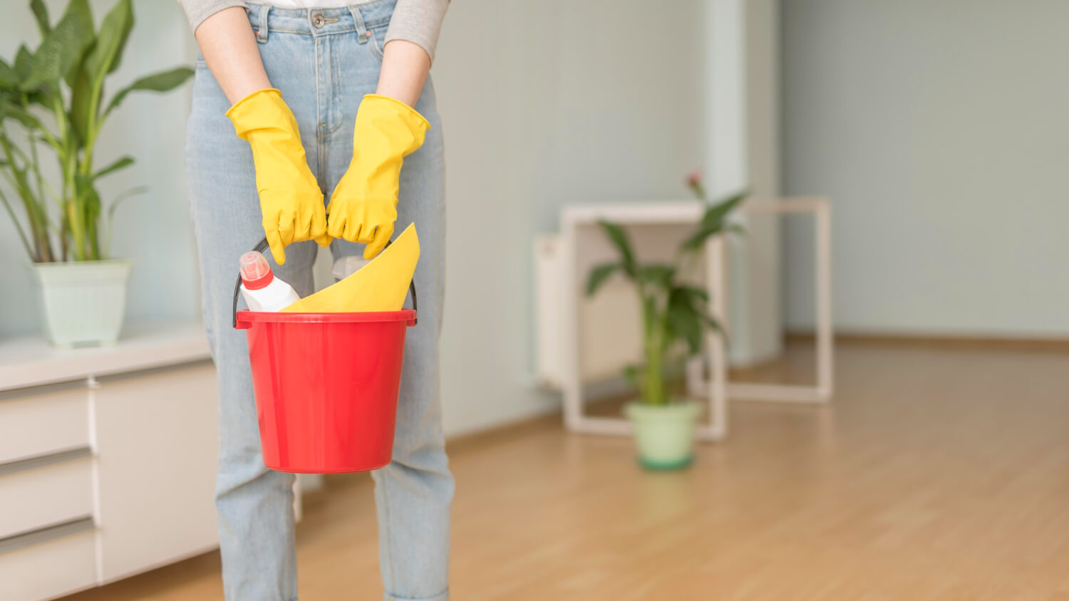 cropped young woman in jeans and yellow protective gloves holding red bucket with cleaning supplies in it