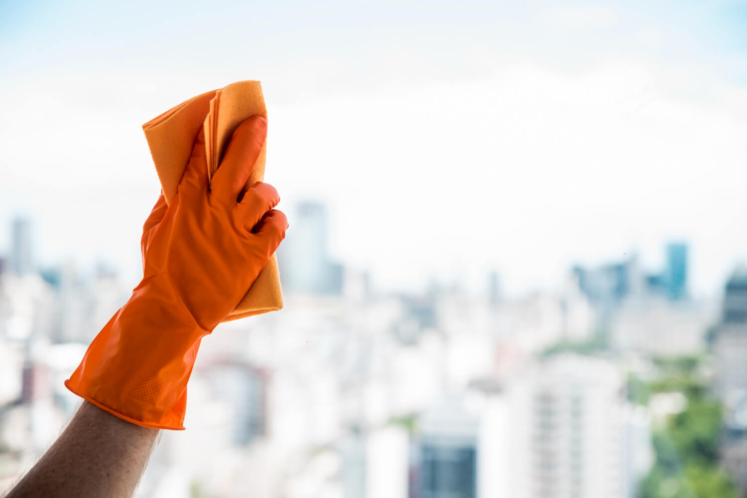 close up male hand with orange glove and orange cloth cleaning window with view of blurred city