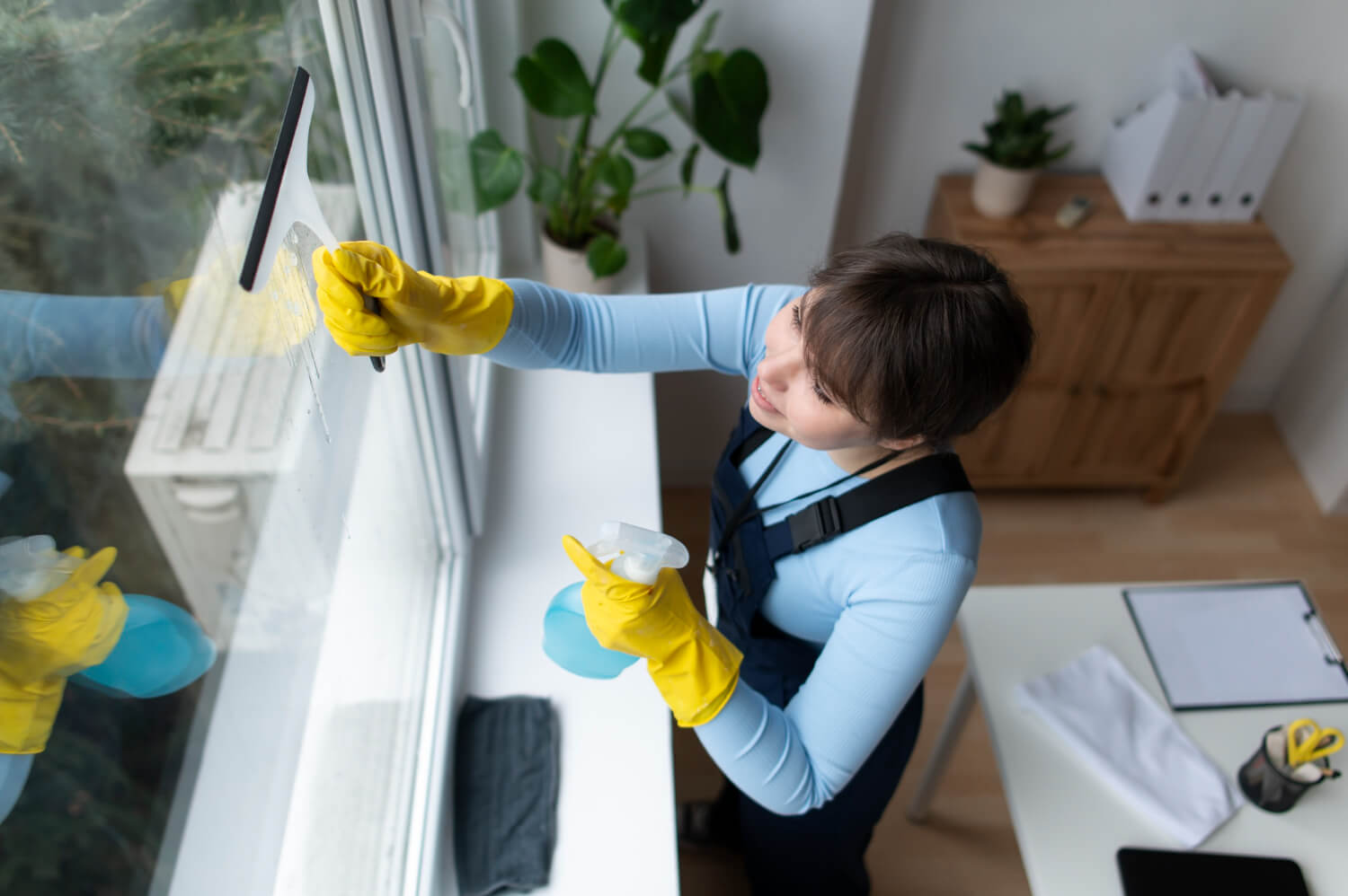 top view of young cleaner in yellow gloves and black uniform cleaning window with spray and cleaning brush