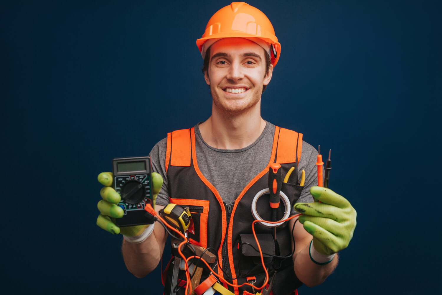 happy electrician smilling at camera while holding digital multimetert in protective gloves
