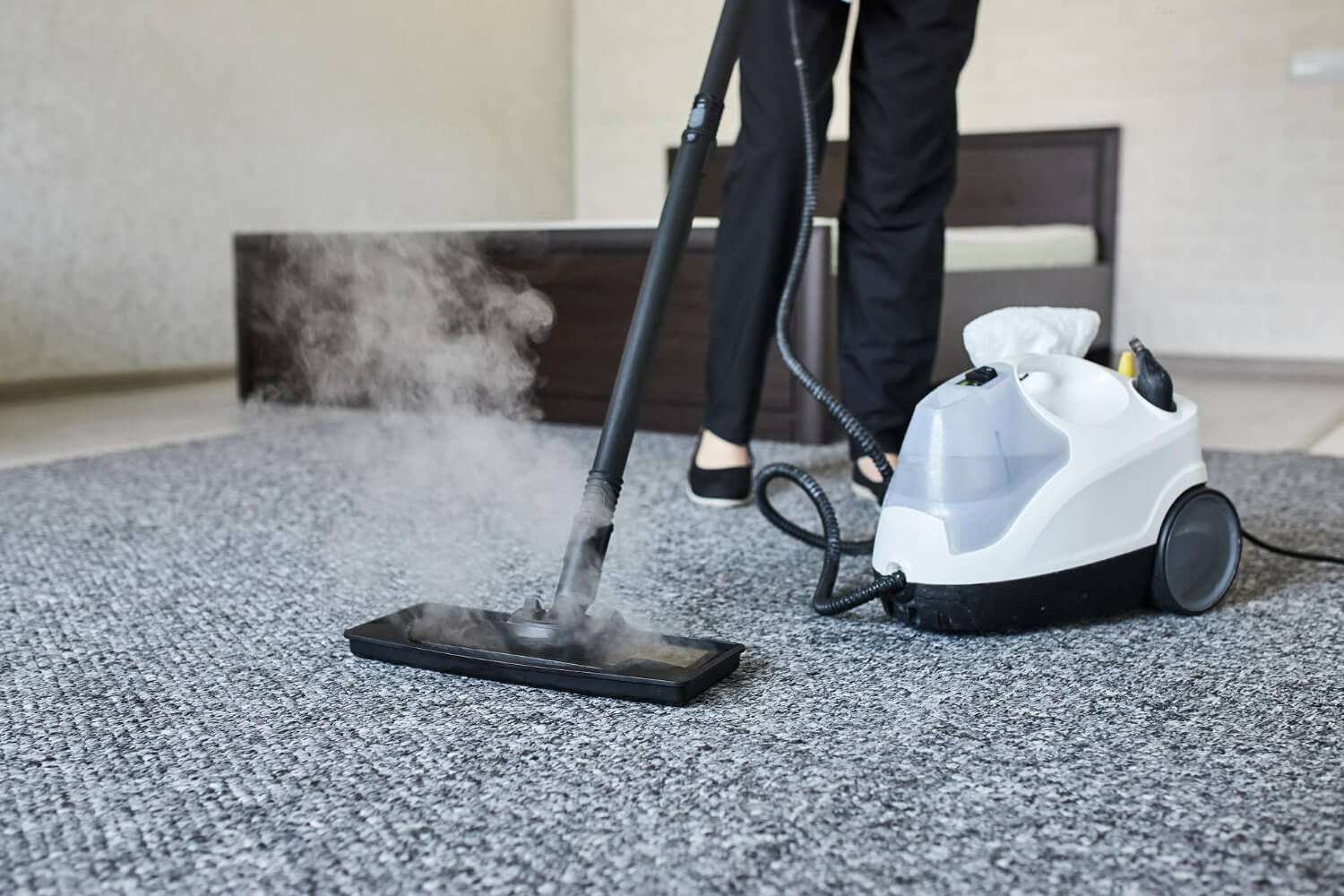 cropped close up of cleaner with black trousers holding professional vacuum while cleaning grey rug in bedroom