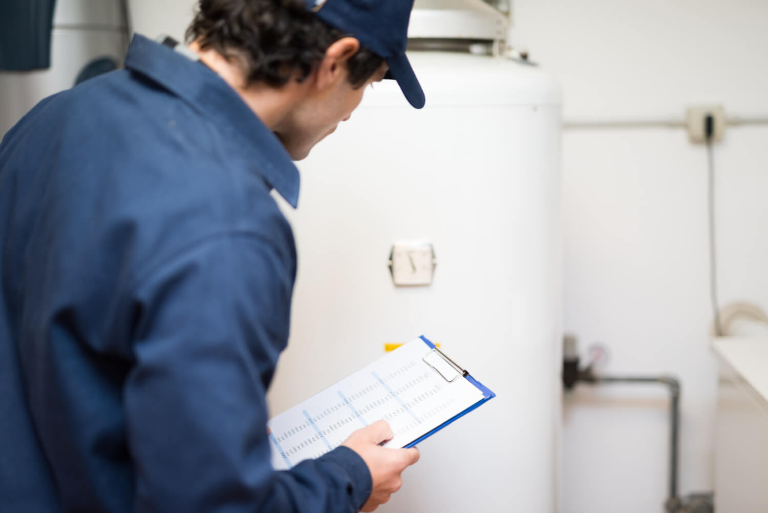 back view of plumber with blue uniform and blue hat looking at boiler while holding notebook