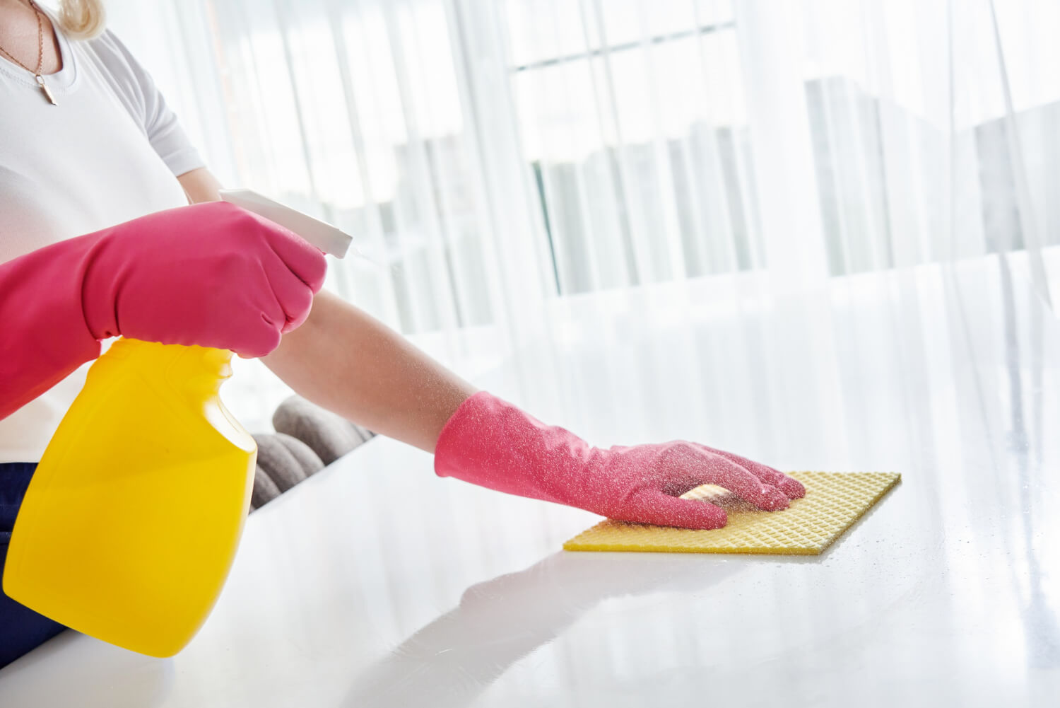 close up cropped woman in pink protective gloves spraying white table and cleaning with yellow cloth