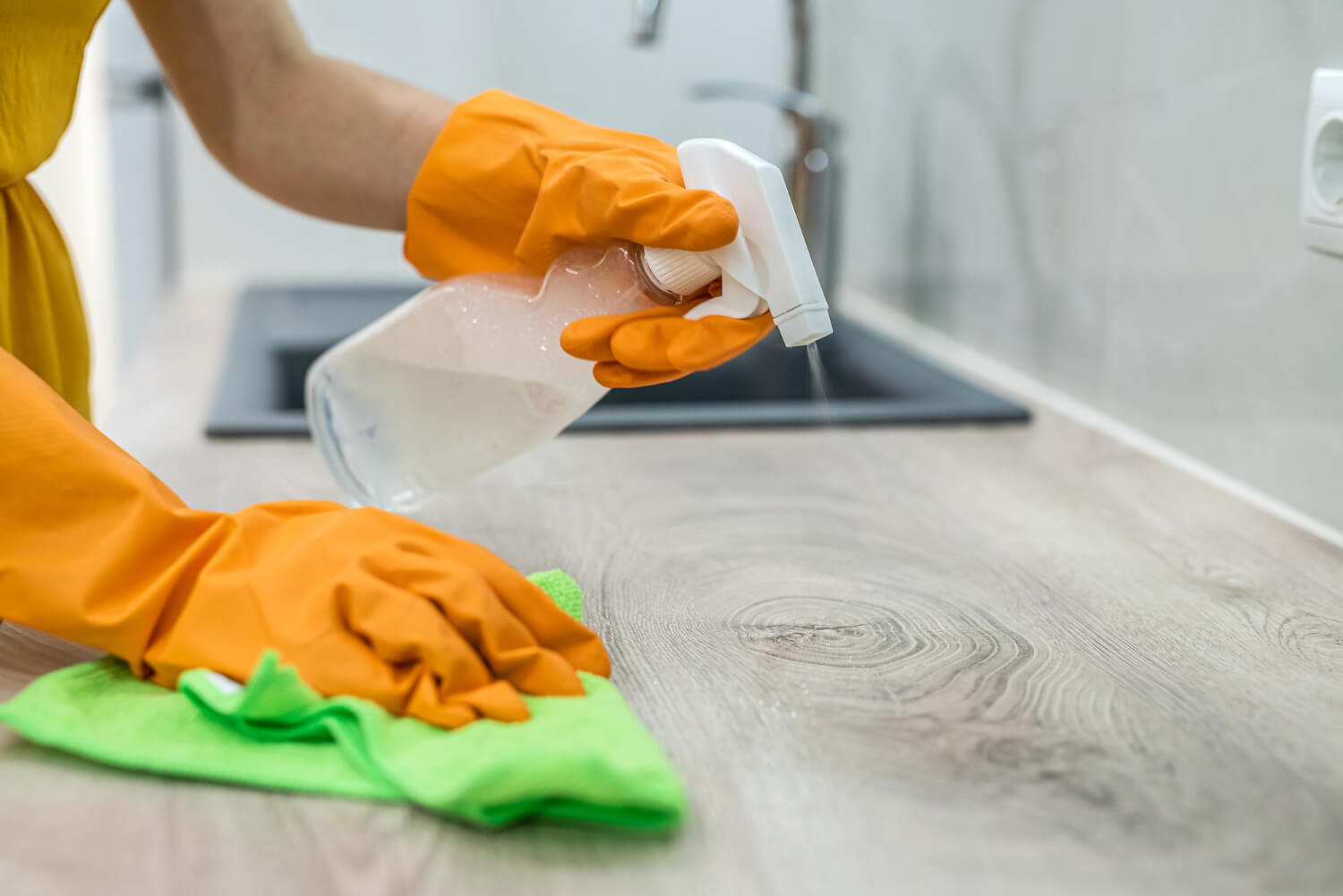 close up female hands in orange protective gloves spraying kitchen and cleaning with green cloth