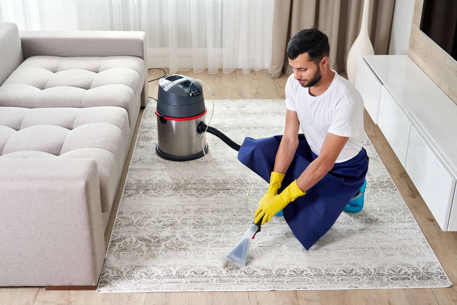 male cleaner in yellow gloves cleaning white carpet with professional vacuum cleaner next to sofa
