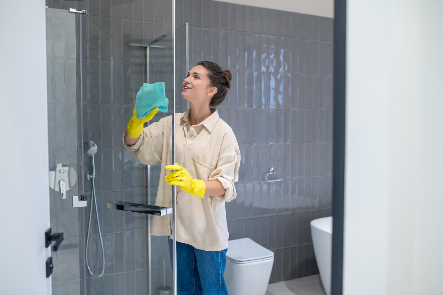 happy woman in shirt and yellow gloves cleaning shower glass with blue cloth