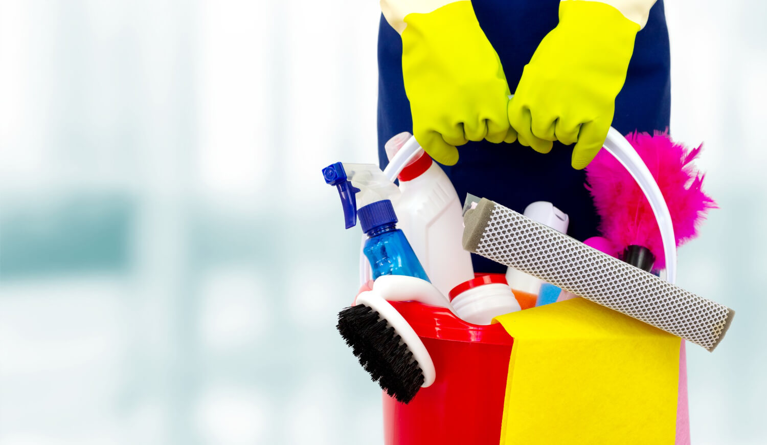 close up female hands in protective gloves holding red bucket with various cleaning products and suplies inside