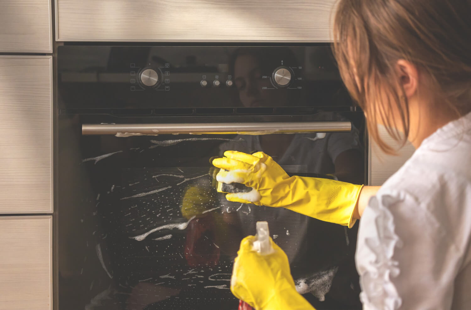 close up back view of dark haired woman in yellow protective gloves cleaning soaped oven door with sponge