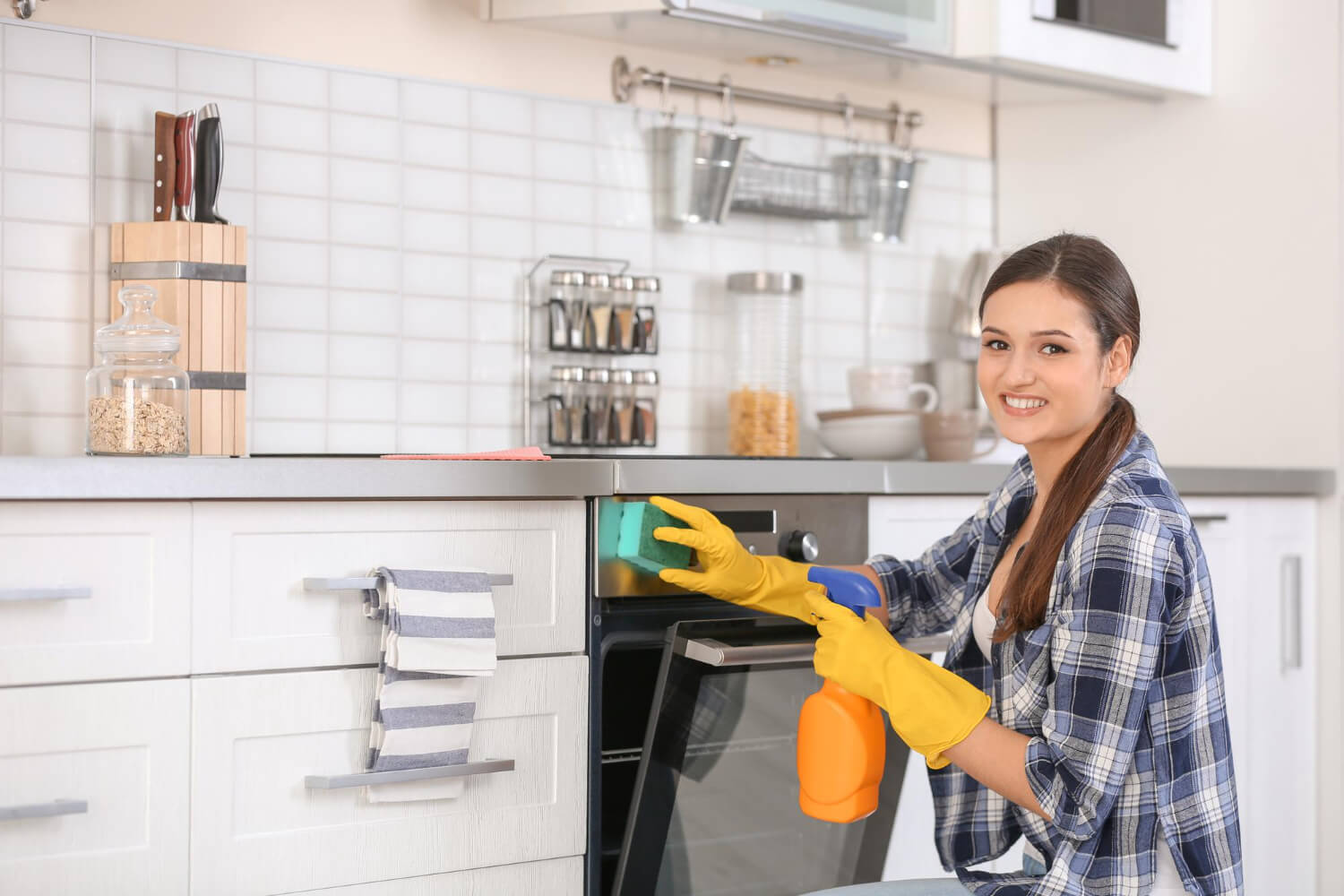 close up happy dark haired woman in yellow gloves cleaning oven with cleaning spray and sponge