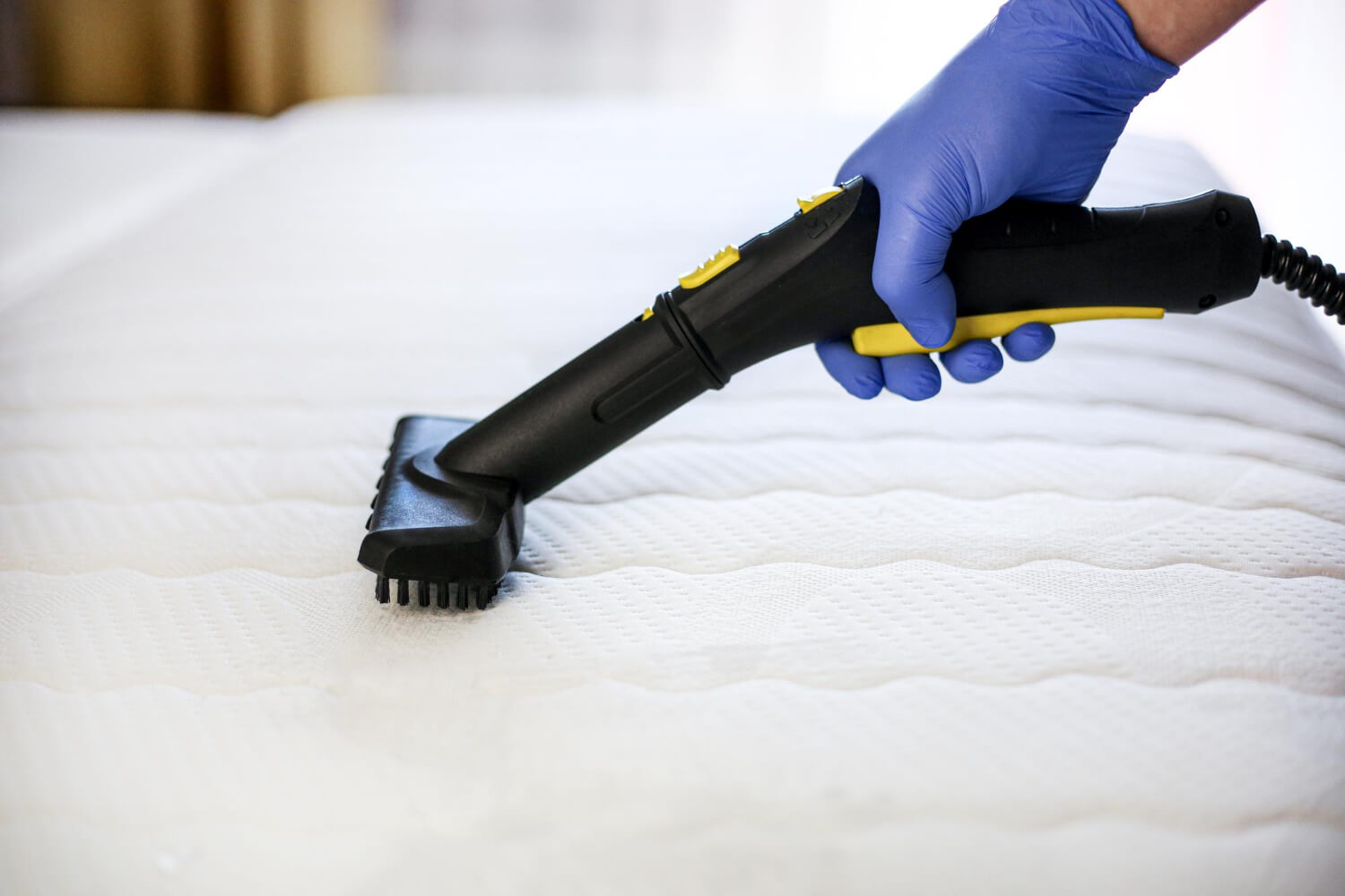 close up cleaner hand in protective blue glove holds small vacuum while cleaning mattress