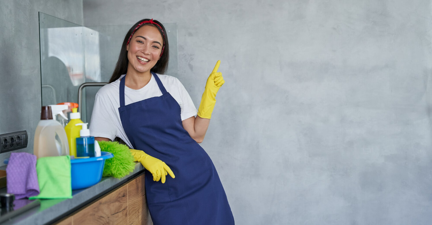 happy young woman smilling at camera while wearing protective yellow gloves and blue uniform with cleaning products next to her