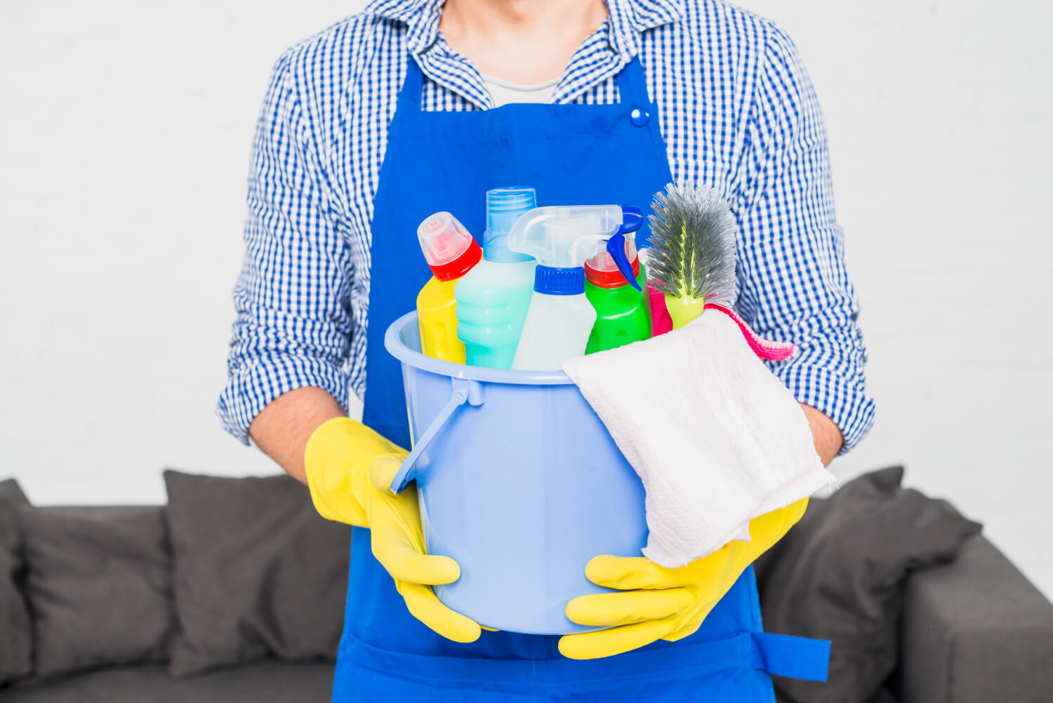 cropped male cleaner in yellow gloves and blue uniform holds grey bucket with cleaning equipment in it