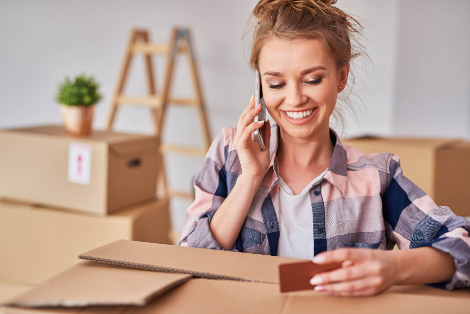 smiling woman using phone while moving house with staffed moving boxes around her