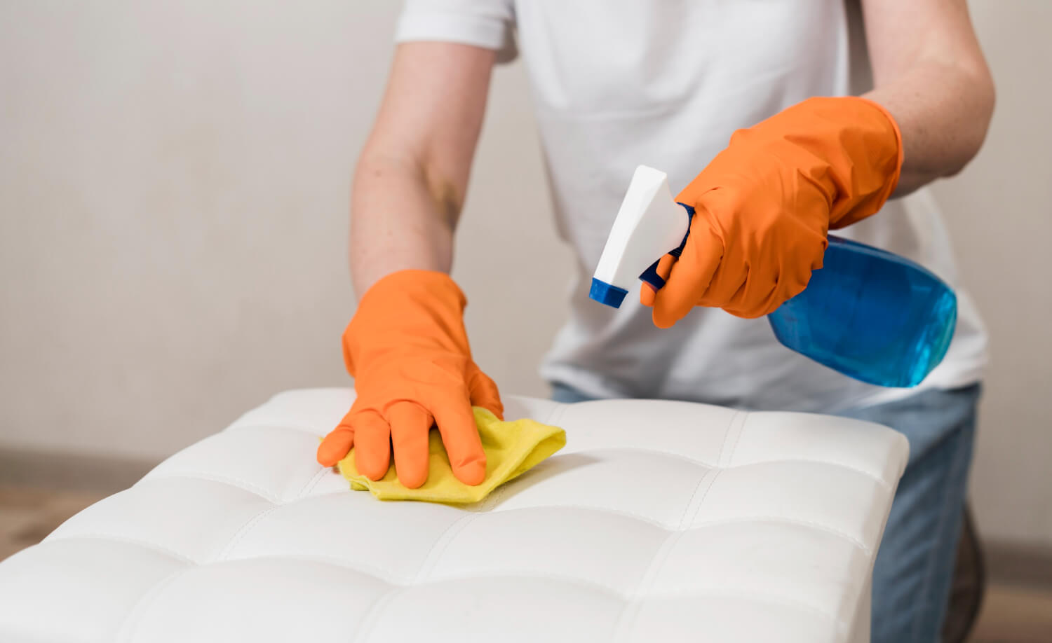 close up male hands in orange gloves cleaning white upholstery with spray and yellow cloth