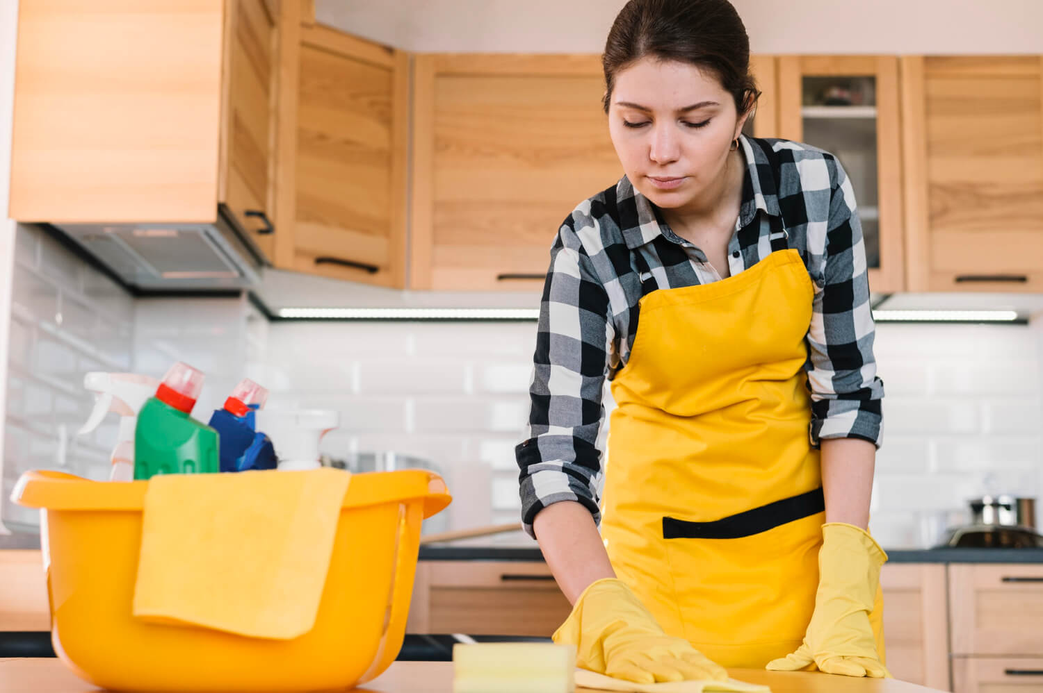 close up female woman in yellow uniform cleaning kitchen table with bucket with cleaning products and supplies next to her