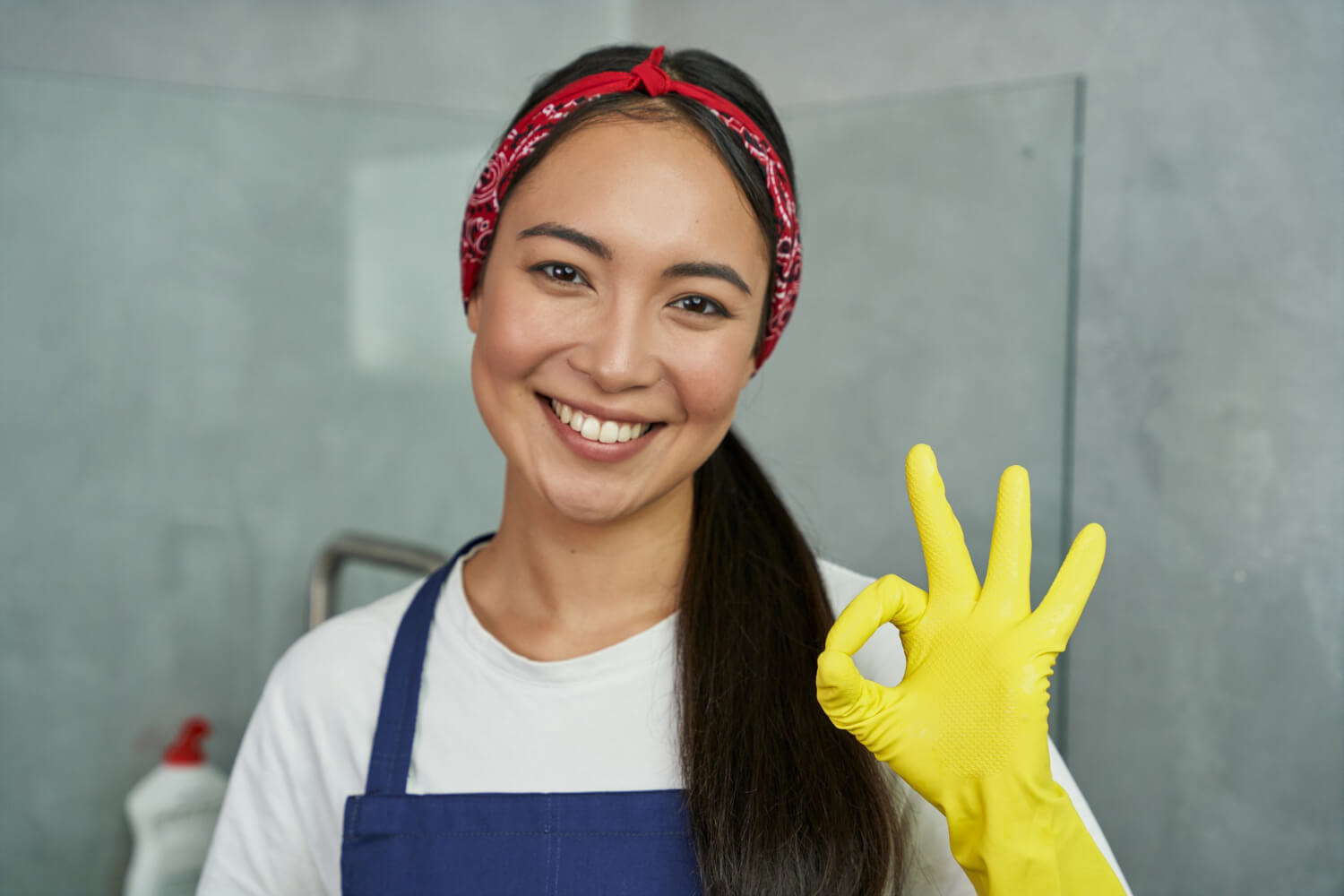 close up happy asian woman in blue uniform red bandana and yellow protective glove smilling at camera while doing ok sign