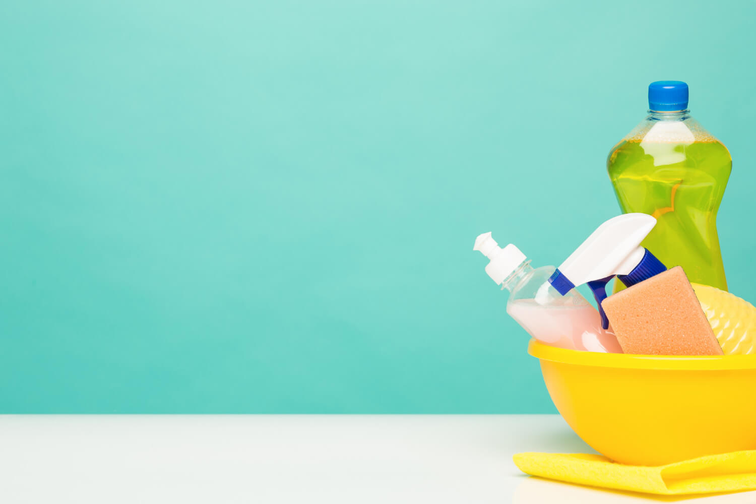 various cleaning bottles and cleaning supplies in yellow bucket with light blue green wall behind