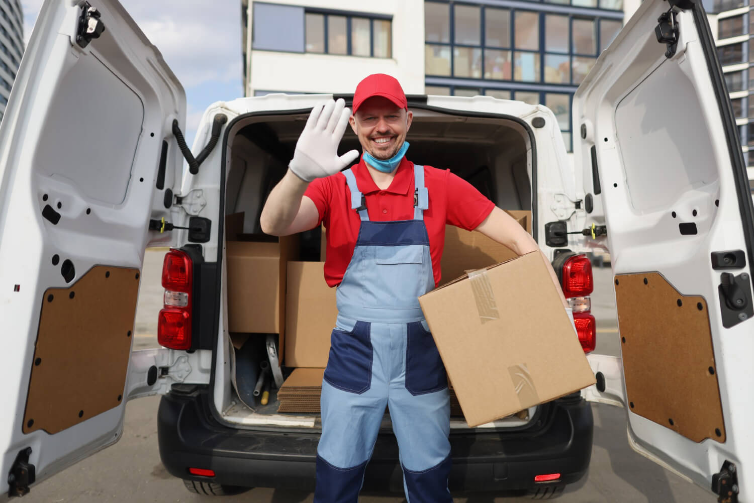 happy mover stands in the back of a moving white van with doors open filled with moving boxes waving at the camera while holding a box