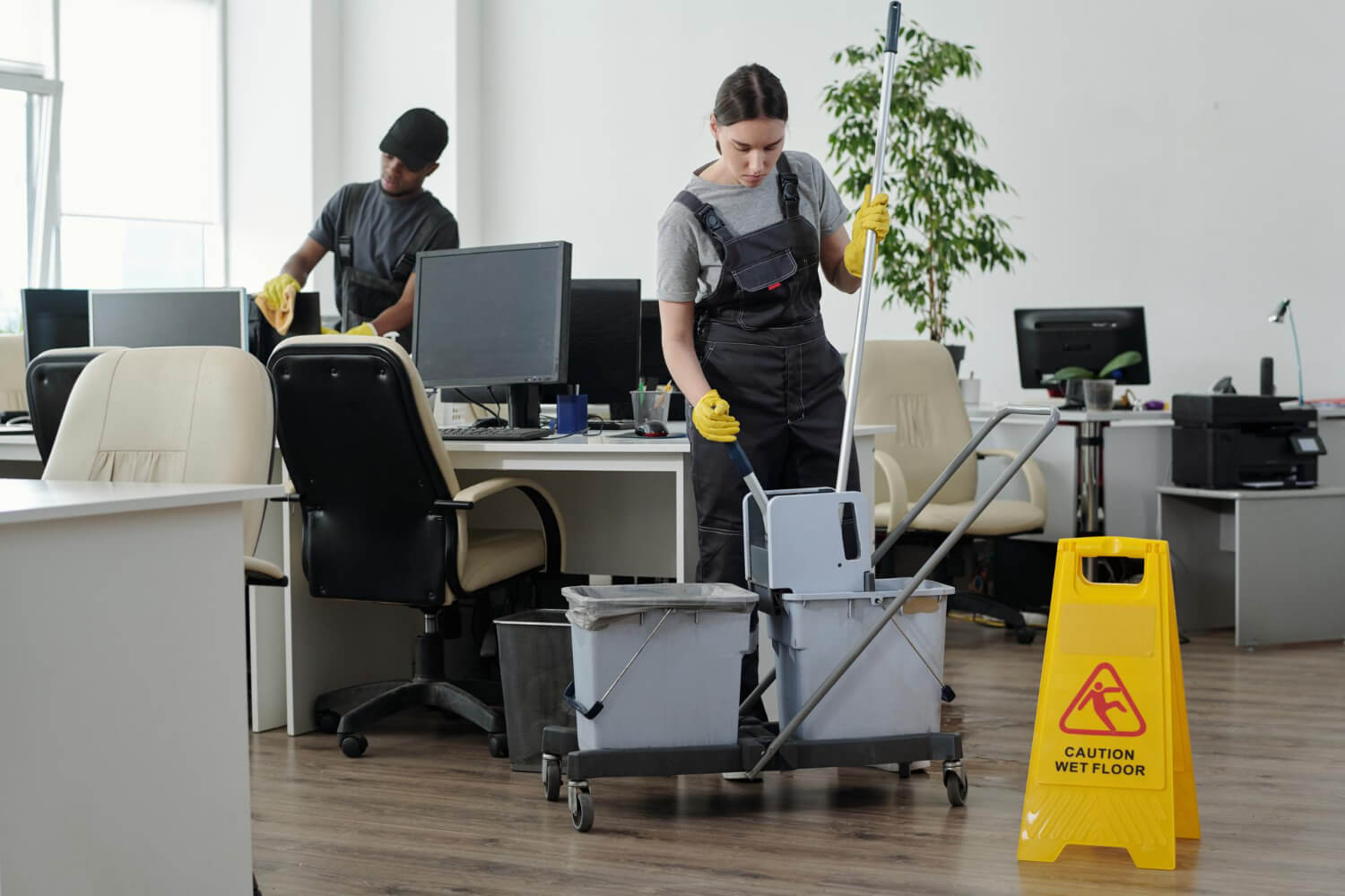 cleaning team in grey uniform and yellow protective gloves cleaning computers and mopping floor in office