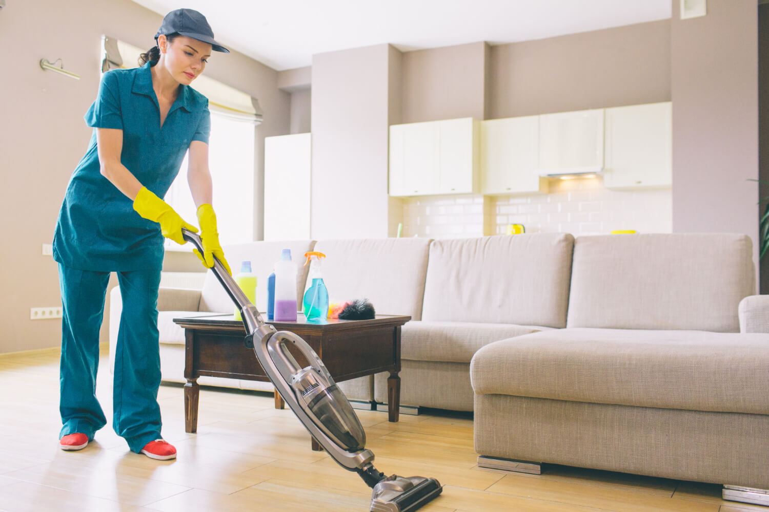 young cleaner in uniform and hat vacuums floor in apartment with big corner sofa